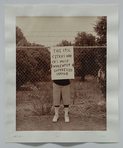 Artist holds sign in front of chainlink fence with citrus trees beyond. Sign says "The 1936 Citrus was OC's most violently suppressed strike"