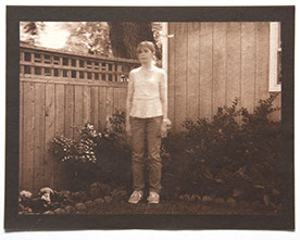 Artist stands center 3/4 view with arms down in corner of backyard. Wood fence, house and plants behind her. All in focus except her upper body 