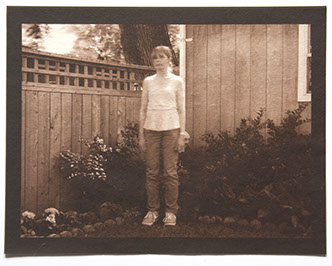 Artist stands center 3/4 view with arms down in corner of backyard. Wood fence, house and plants behind her. All in focus except her upper body 