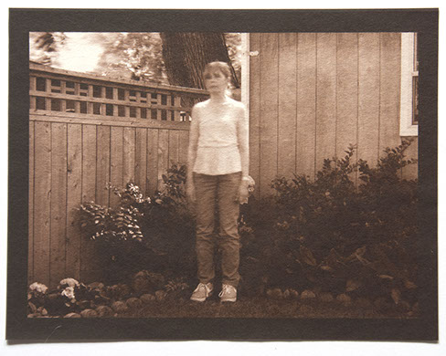 Artist stands center 3/4 view with arms down in corner of backyard. Wood fence, house and plants behind her. All in focus except her upper body 