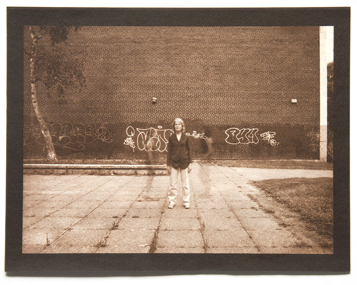 Artist stands ctr facing forward. Large brick wall with graffiti behind. Square pavers with weeds form a grid below. 2 ghost figures flank her.