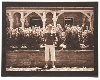 Artist stands ctr face forward in bright day. Behind her a low wall /w small shrubs reaches her height. Backgrnd is an ornate wood & stone porch