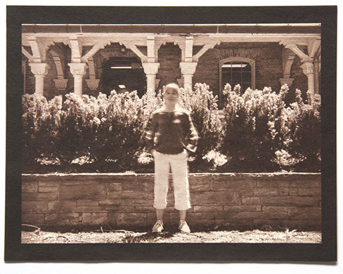 Artist stands ctr face forward in bright day. Behind her a low wall /w small shrubs reaches her height. Backgrnd is an ornate wood & stone porch