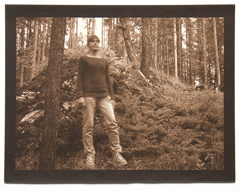 Artist stands off ctr at 3/4 profile facing right at base of shrubby mound which reaches her head. Tree trunks surround her.