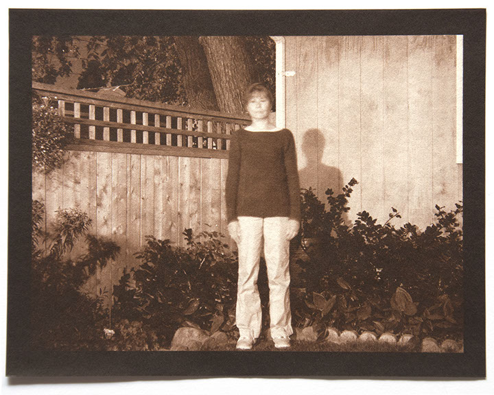 Artist stands in same position in backyard surround by wood fence, house, shrubs. It is nighttime. Brightly lit /w her shadow on wall behind her