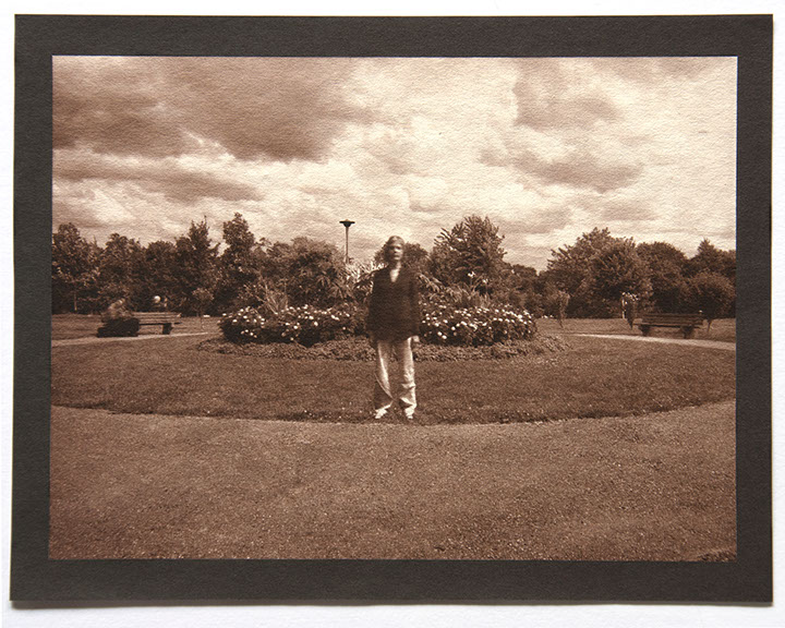 Artist stands ctr facing forward in round landscape of grass. Flowering shrubs at center behind her. Rolling clouds above and trees in distance.