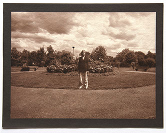 Artist stands ctr facing forward in round landscape of grass. Flowering shrubs at center behind her. Rolling clouds above and trees in distance.