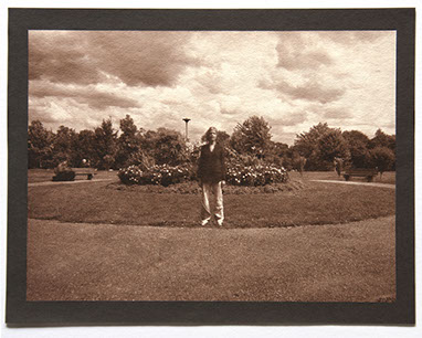 Artist stands ctr facing forward in round landscape of grass. Flowering shrubs at center behind her. Rolling clouds above and trees in distance.