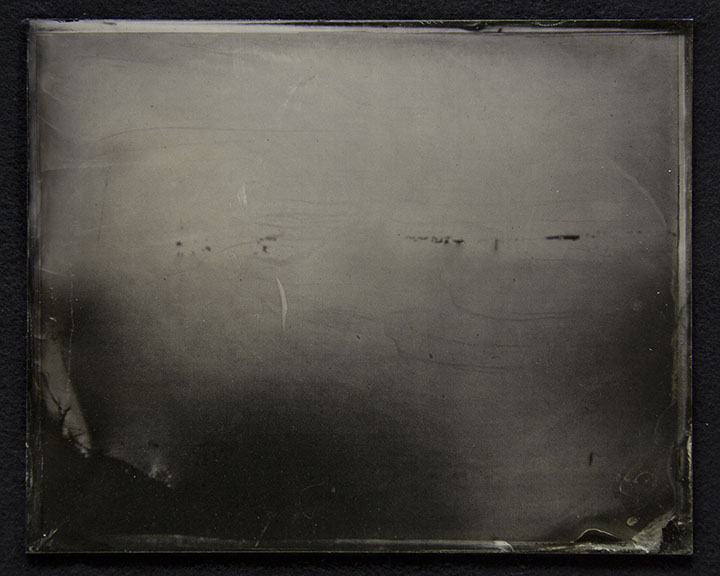 C3: Landscape photo on glassplate. Medium cool greys above-Dark warm grey below. Small dark scrapes as image or photochemical residue. U decide.