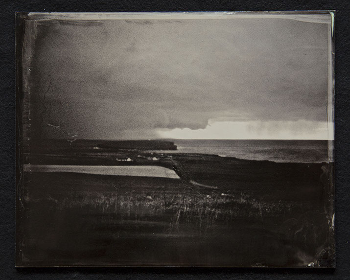 Panel C2-Land, grasses & shoreline dominate bottom half. Houses near the shore. Sky heavy with clouds with 1 clear sliver over ocean in distance