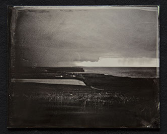 Panel C2-Land, grasses & shoreline dominate bottom half. Houses near the shore. Sky heavy with clouds with 1 clear sliver over ocean in distance