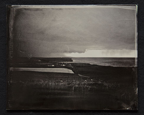 Panel C2-Land, grasses & shoreline dominate bottom half. Houses near the shore. Sky heavy with clouds with 1 clear sliver over ocean in distance
