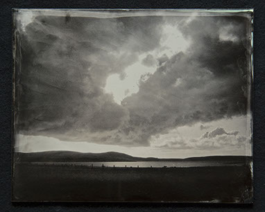 Panel C3 - Big sky full of dark clouds. Light peaks through and below. A low horizon of landscape with a lake surrounded by land and hills.